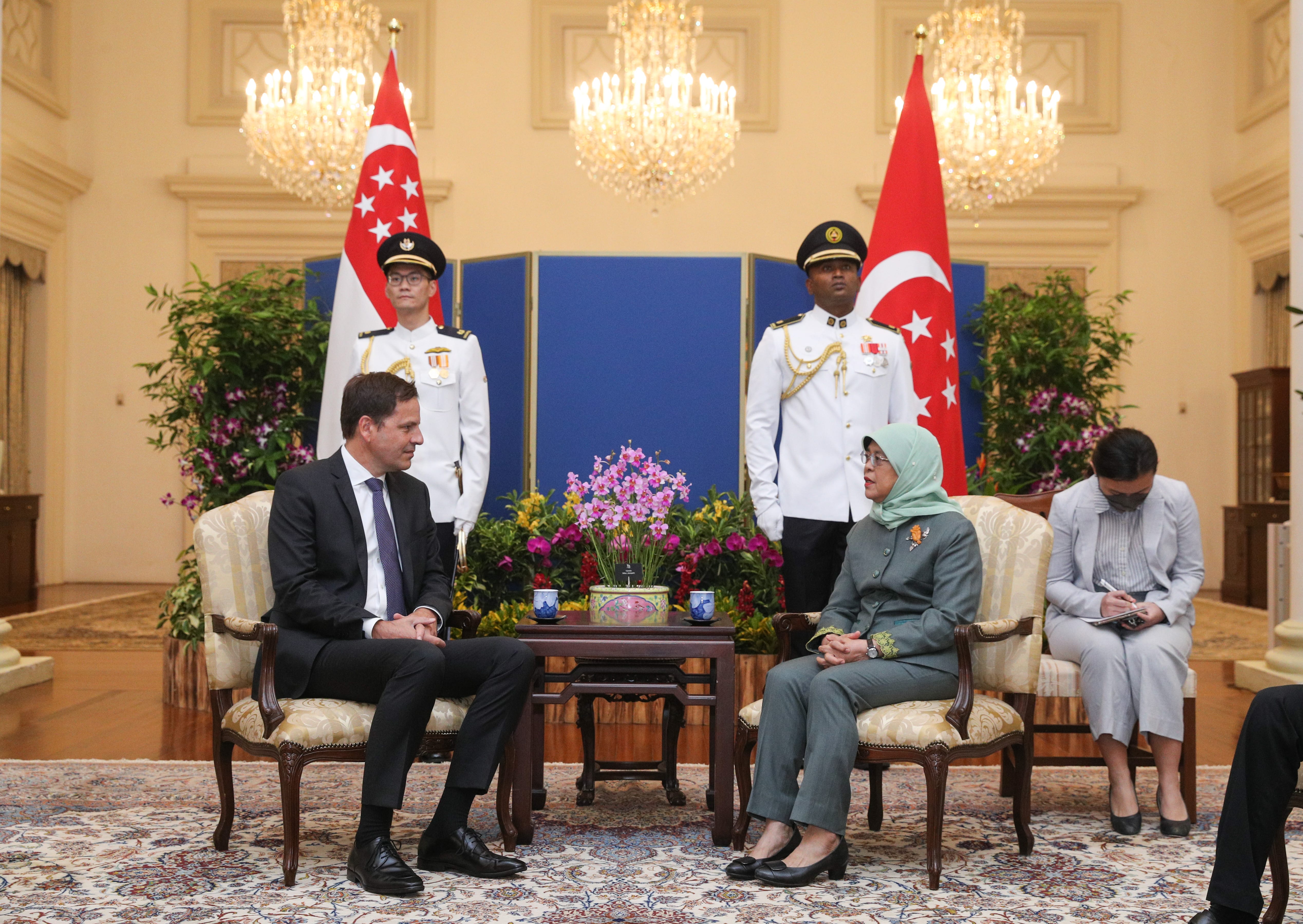 Man in suit and Halimah Yacob seated facing each other, guards and Singapore flags in background.